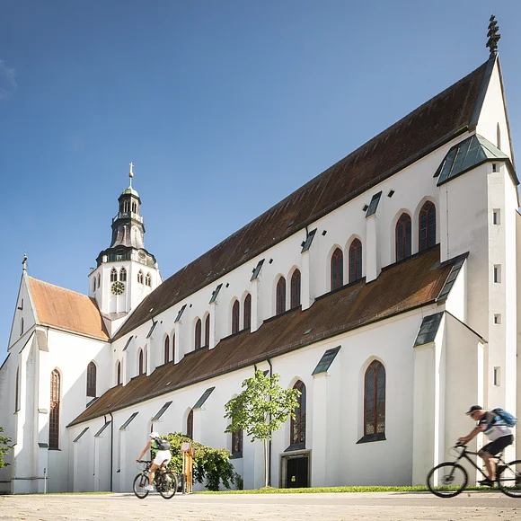 Weißes Kirchgebäude mit Turm und zwei Radfahrern im Vordergrund bei blauem Himmel.