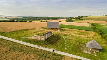 Vier Radfahrer fahren auf einem Schotterweg neben einem rekonstruierten keltischen Fürstensitz.
