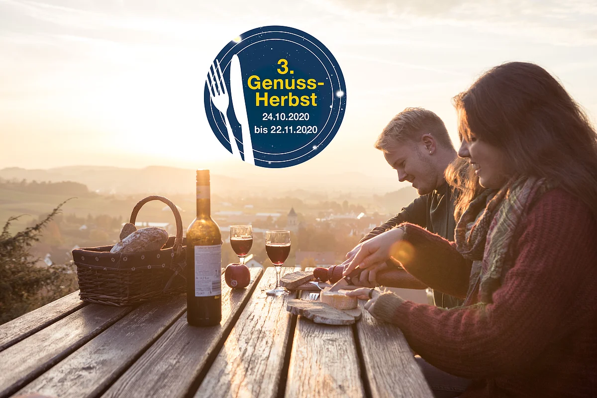 Eine Frau und ein Mann sitzen an einem mit Brotzeit und Wein gedecktem Holztisch. In der Mitte ist das Logo des "3. Genussherbst - 24.10.2020 bis 22.11.2020" eingeblendet.