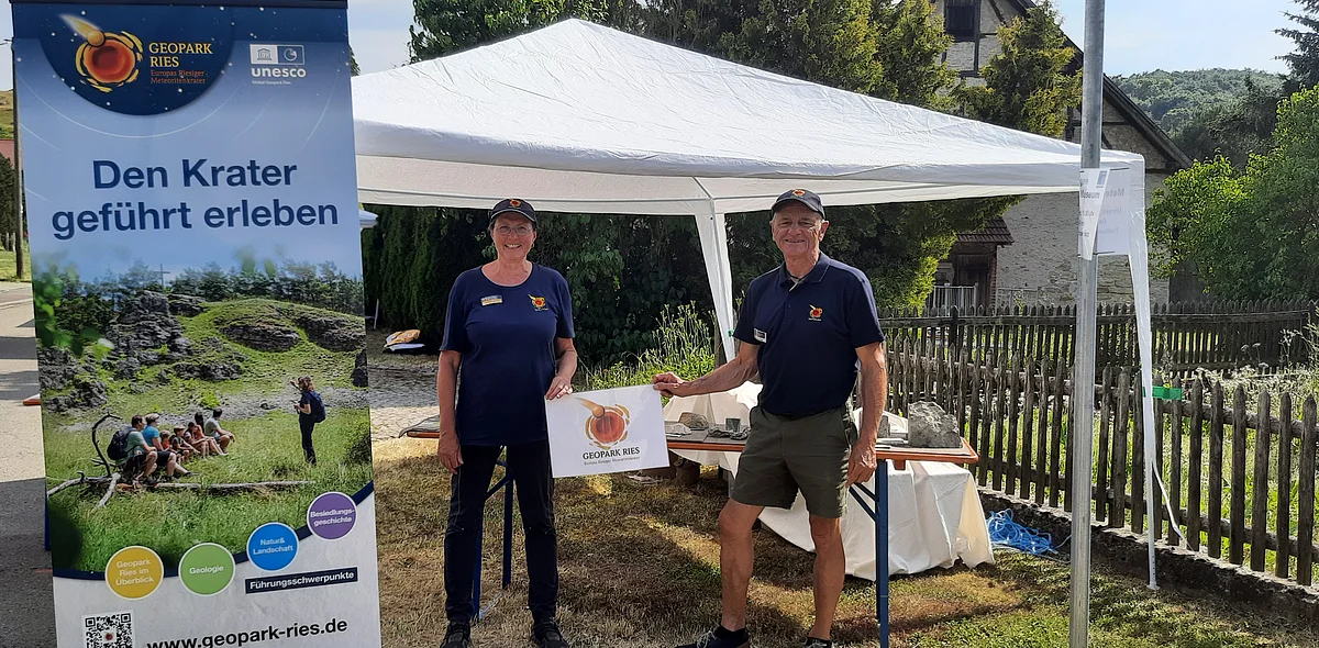 Zwei Geoparkführer:innen stehen neben einem Poster mit der Aufschrift "Den Krater geführt erleben" unter einem weißen Pavillon und halten ein Schild mit dem Logo des Geoparks Ries.
