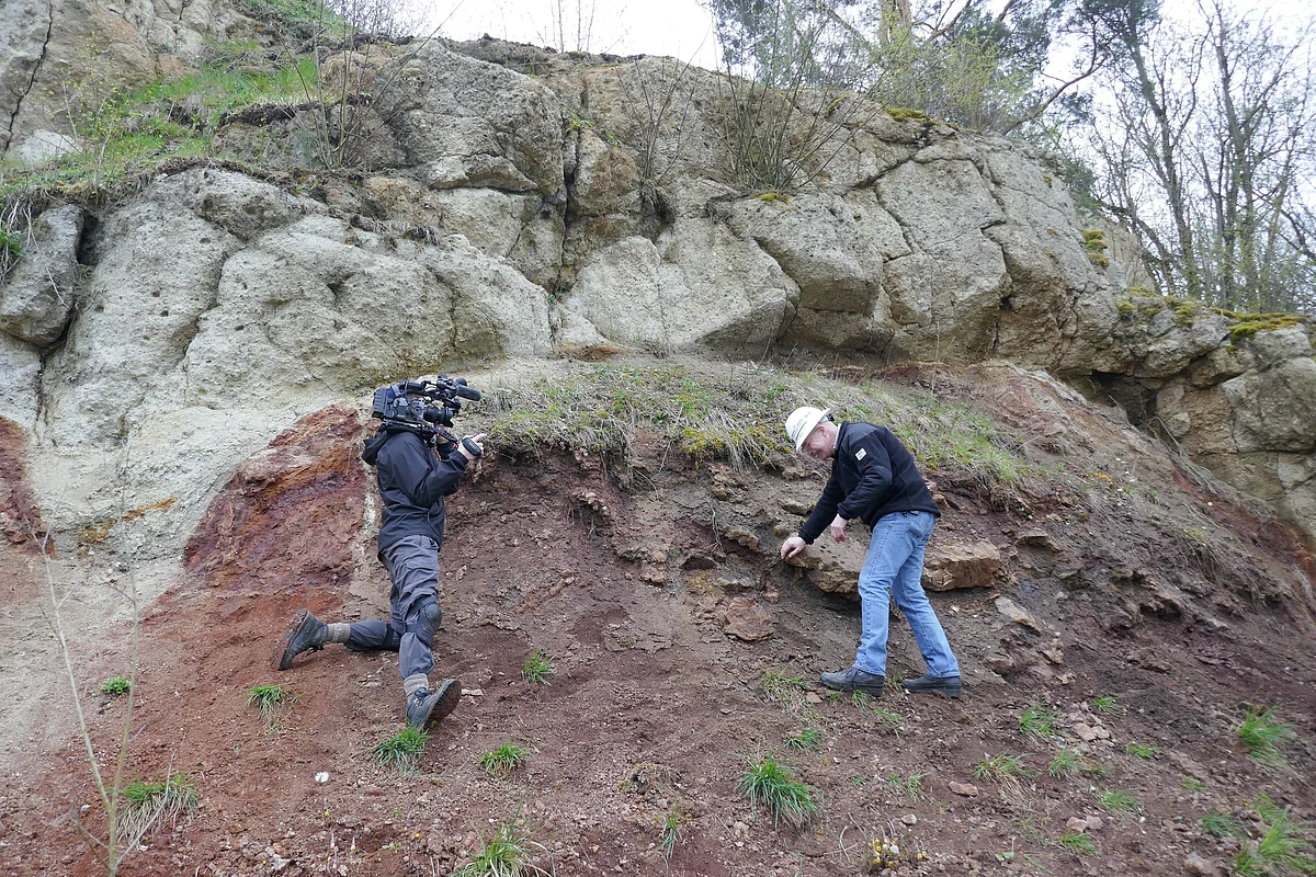 BBC mit Prof. Dr. Harald Hiesinger An einem steilen Hang unterhalb einer Felsformation stehen ein Mann mit Fernsehkamera und ein Mann mit Schutzhelm.