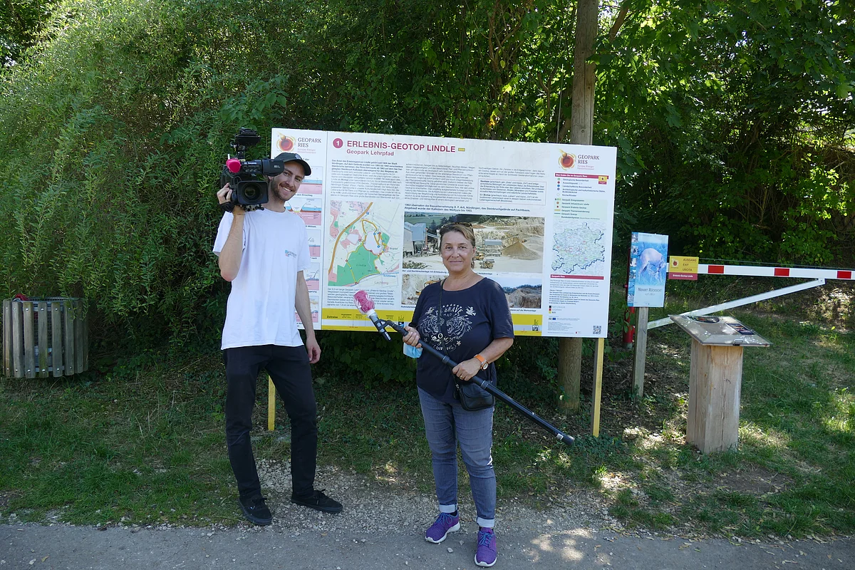 Zwei Personen mit Filmequipment stehen vor einer großen Ereignistafel mit dem Titel "Erlebnis-Geotop Lindle".