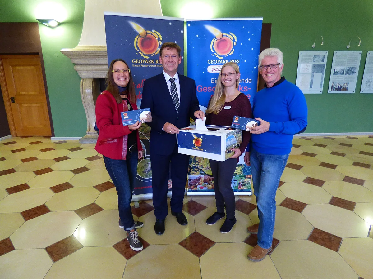 In einem gefliesten Raum mit grünen Wänden stehen Heike Burkhardt, Stefan Rößle und Günther Zwerger vor Plakaten mit dem Logo des Geopark Ries. Eine weitere Person hält eine große Box mit dem Logo des Geopark Ries.