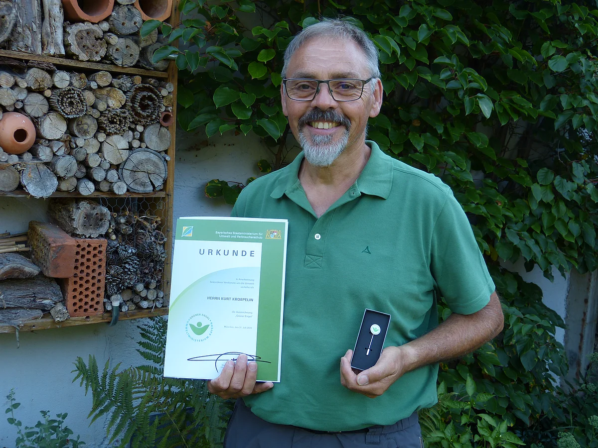 Ein Mann in einem grünen Poloshirt hält eine Urkunde in den Händen. An der Hauswand hinter ihm ist ein Insektenhotel und Blätter zu sehen.