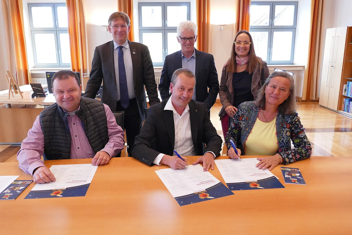 Drei Personen sitzen an einem Tisch und unterzeichnen jeweils eine Urkunde. Dahinter stehen Stefan Rößle, Günther Zwerger und Heike Burkhardt.