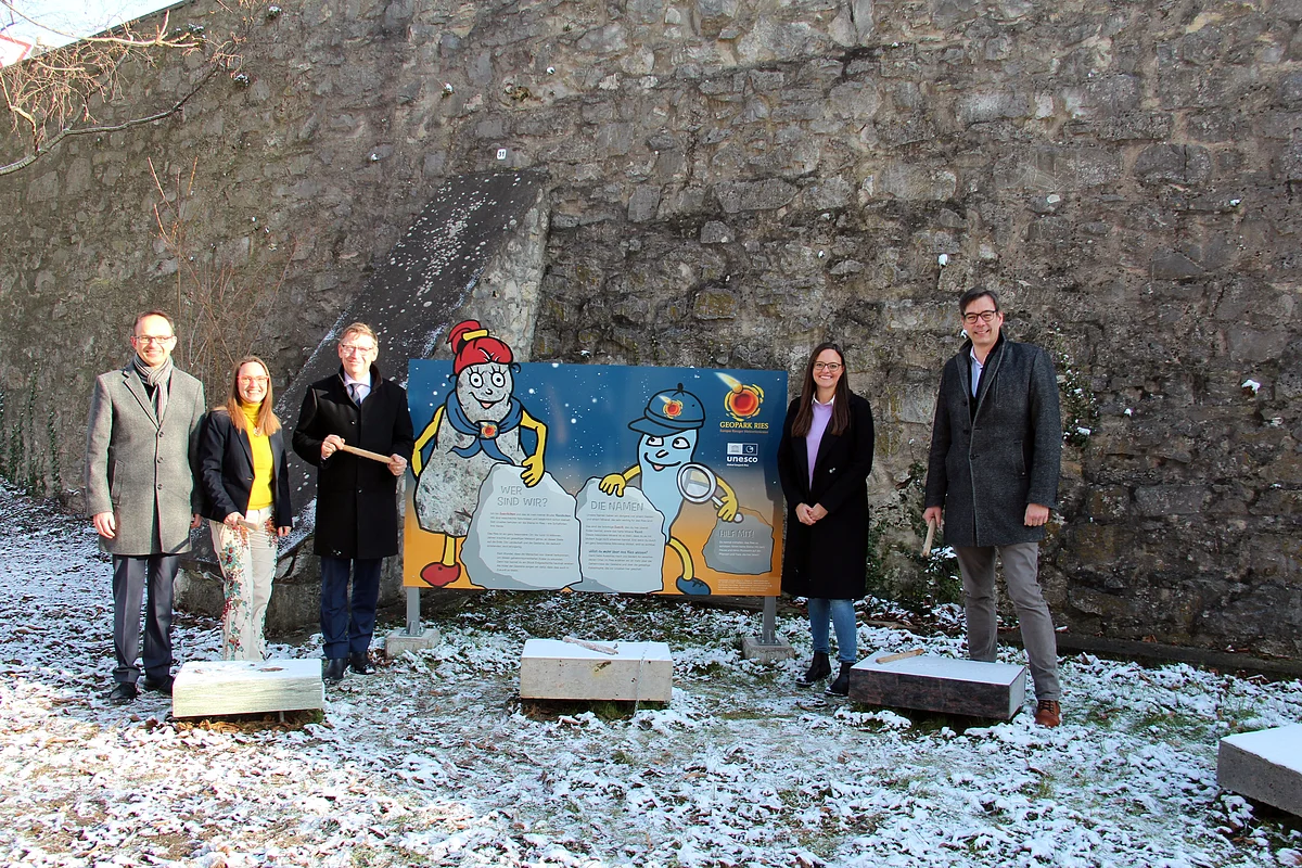 Vor einer hohen Mauer steht eine Informationstafel mit den Maskottchen Suevitchen und Riesitchen, daneben stehen einige Personen. Auf dem Boden liegen geschliffene Steinquader.
