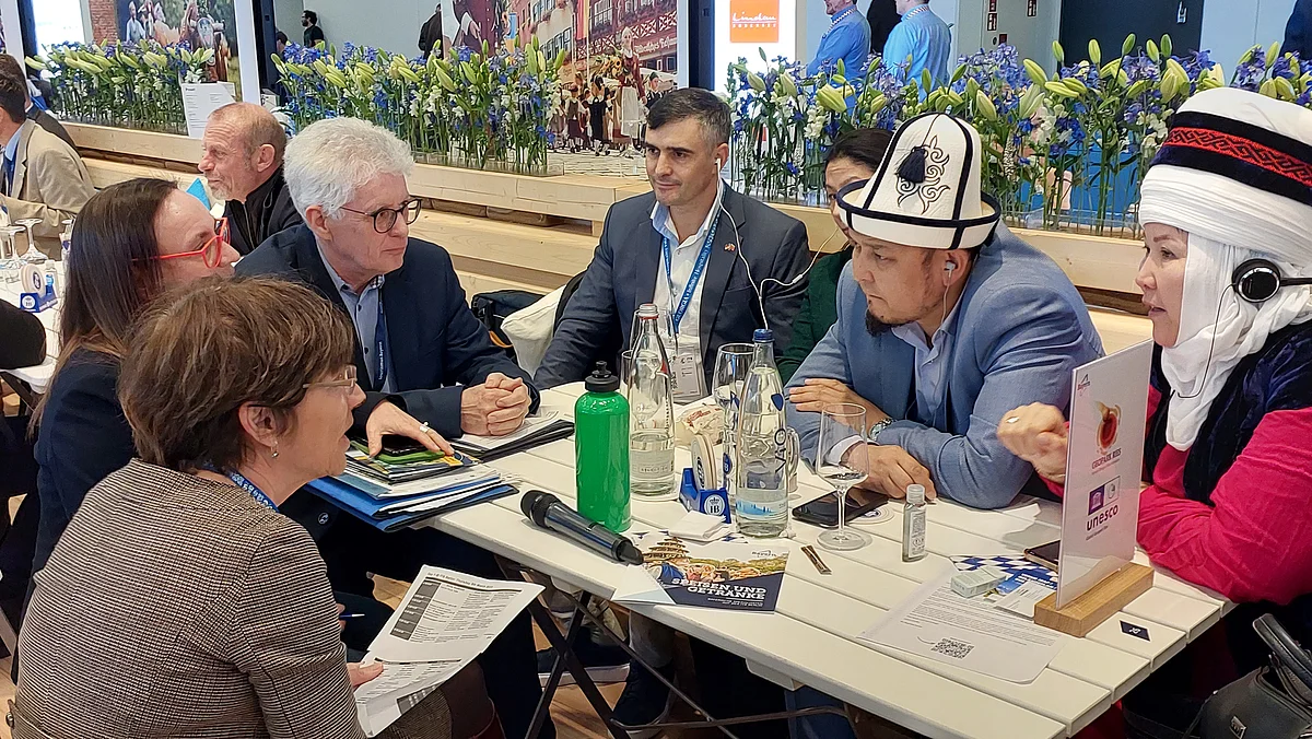 Auf einem Tisch stehen Getränke und liegen Unterlagen, darum sitzen mehrere Personen internationaler Herkunft bei einer Besprechung.