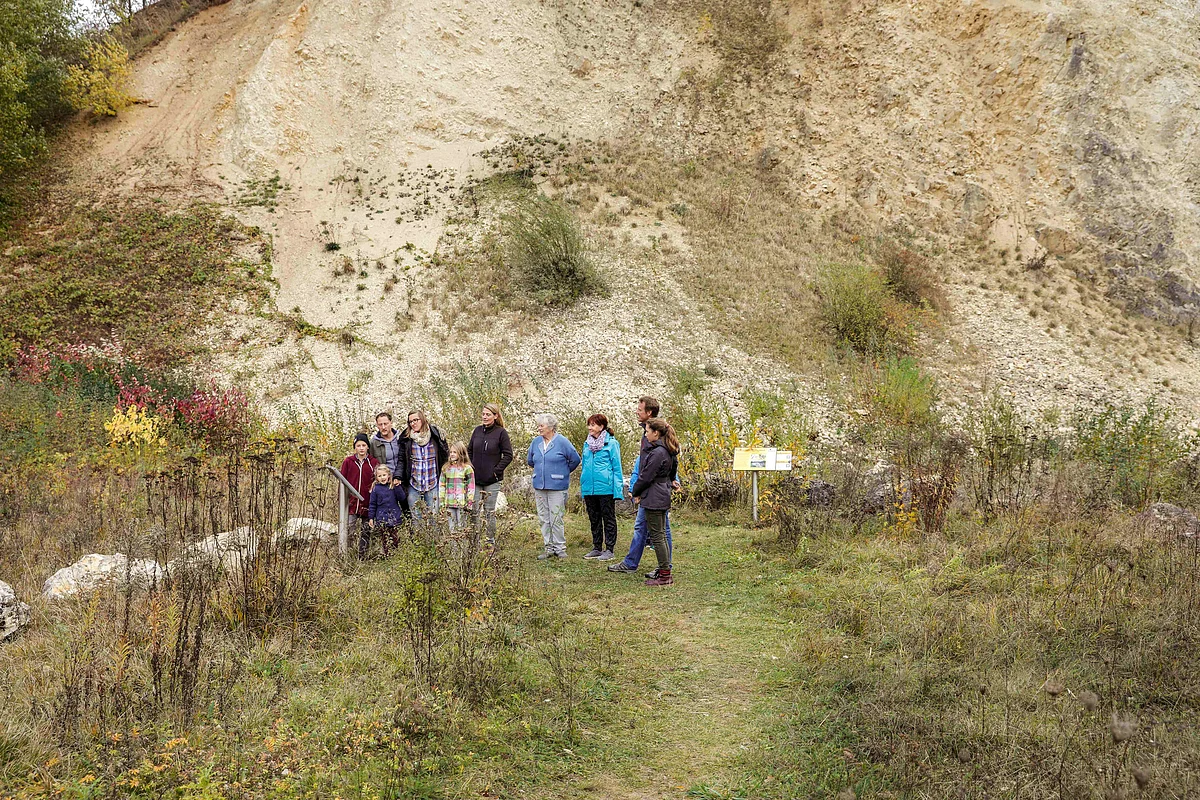 Eine Gruppe Personen steht auf einer herbstlichen Wiese vor einer kleinen Informationstafel. Im Hintergrund ist eine Felswand zu sehen.