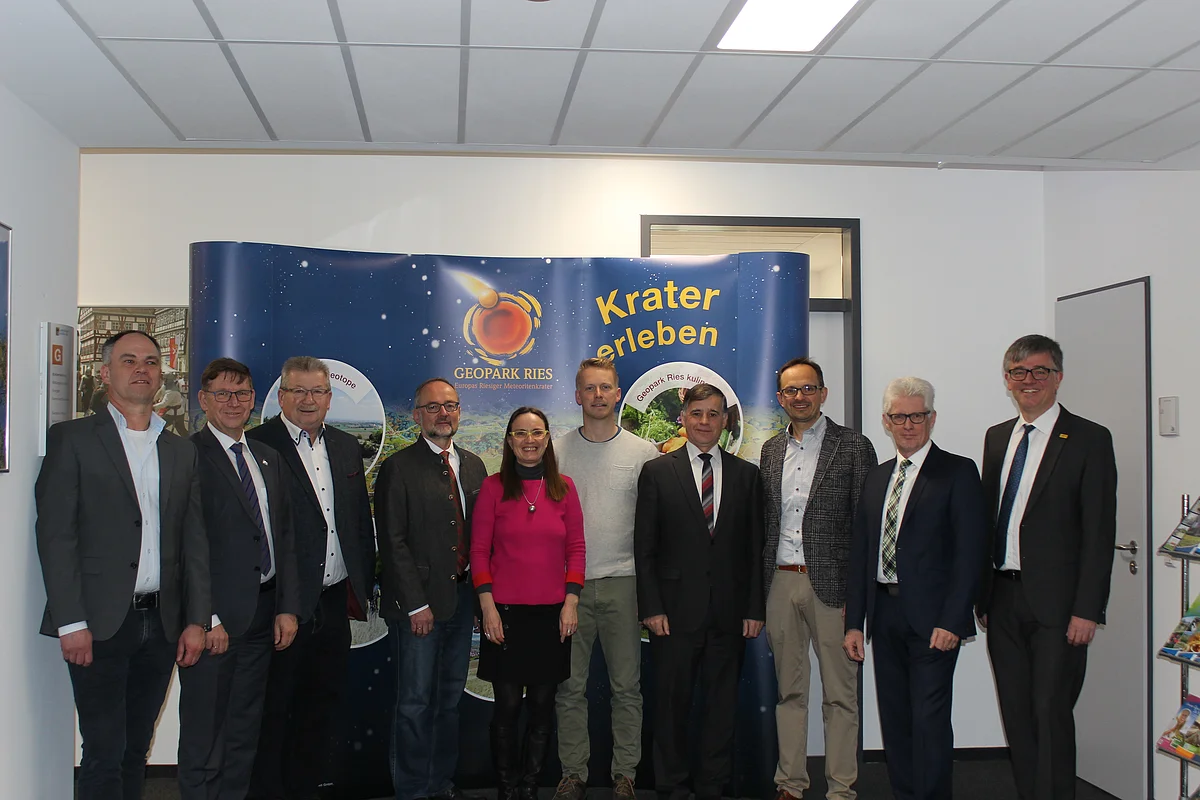 Eine Gruppe Personen steht vor einer dunkelblauen Stellwand mit dem Logo des Geopark Ries und dem Schriftzug "Krater erleben".