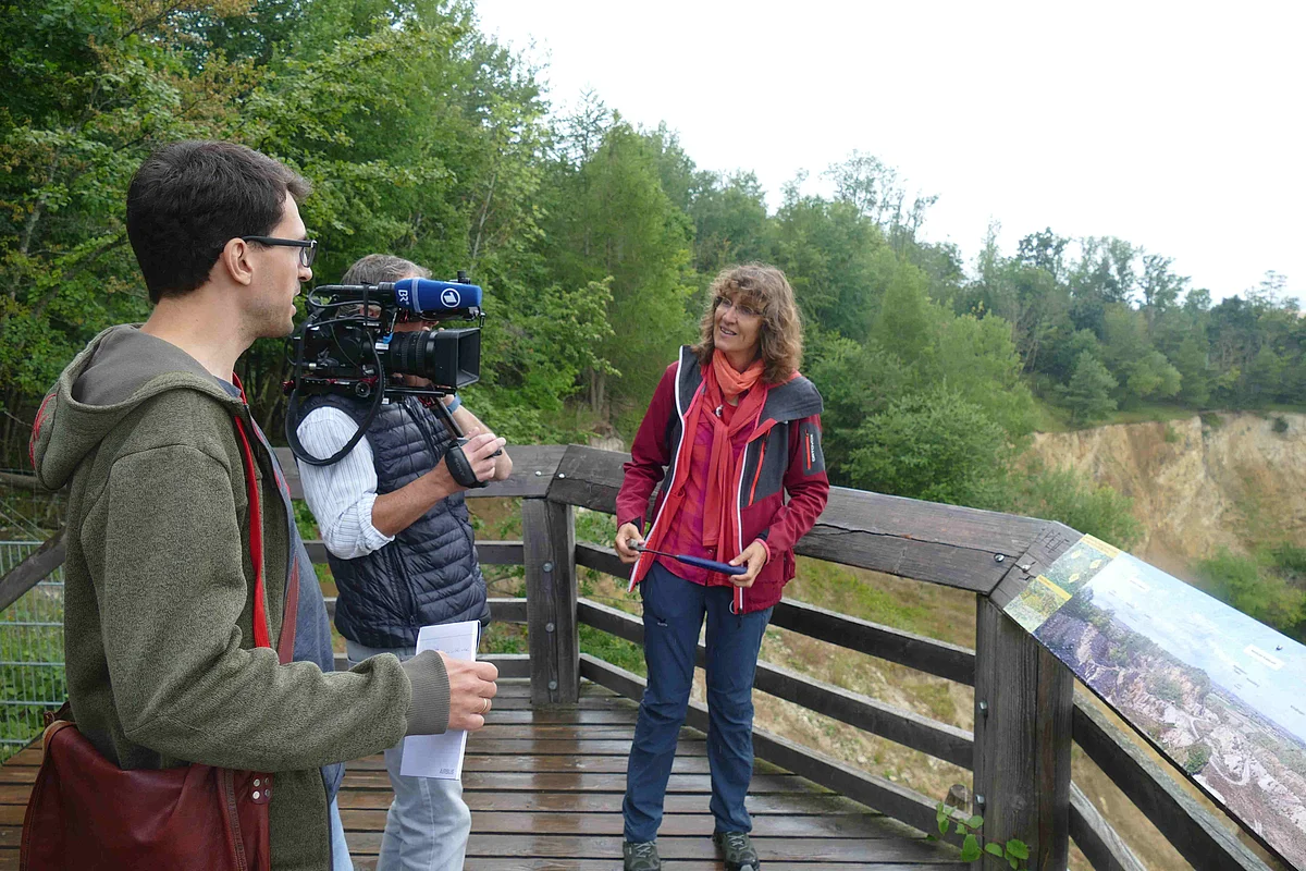 Zwei Personen mit Filmequipment stehen mit einer Frau auf einer Holzplattform oberhalb eines ehemaligen Steinbruchs.