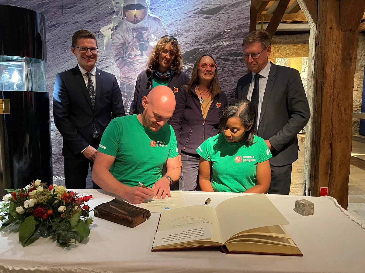 Zwei Personen in grünen T-Shirts unterschreiben in einem großen Buch. Im Hintergrund ist ein Bild eines Astronauten zu sehen, vor dem weitere Personen stehen.