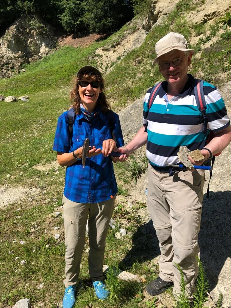 In einem ehemaligen Steinbruch steht ein Mann mit Gesteinsbruchstücken in der Hand und gibt einer Frau einen Geologenhammer.