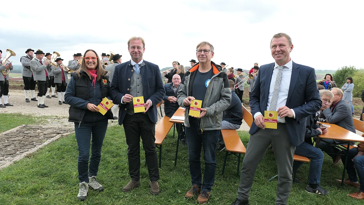 Vier Personen halten Wanderwegsschilder in den Händen. Im Hintergrund sind weitere Personen auf Bierbankgarnituren zu sehen.