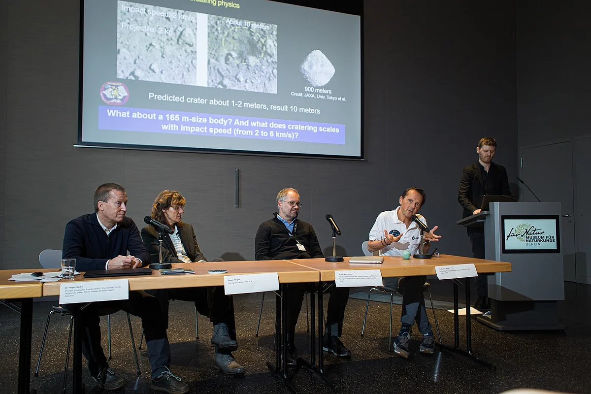 An einem Tisch mit Namensschildern und Mikrofonen sitzen vier Personen. An der Wand dahinter ist eine Leinwand mit Präsentationsfolien zu sehen.