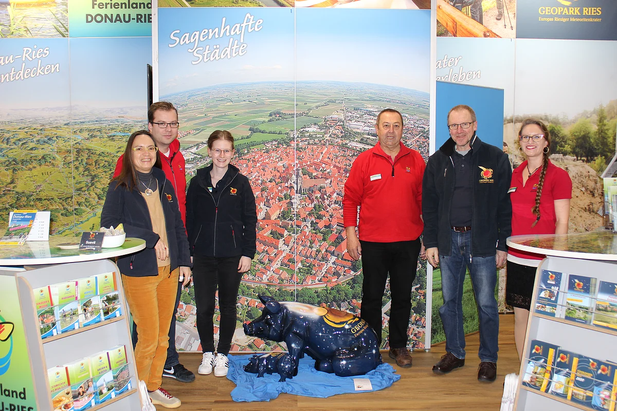 Sechs Personen in dunkelblauen oder roten Oberteilen stehen an einem Messestand mit Aufstellern für Informationsflyer.