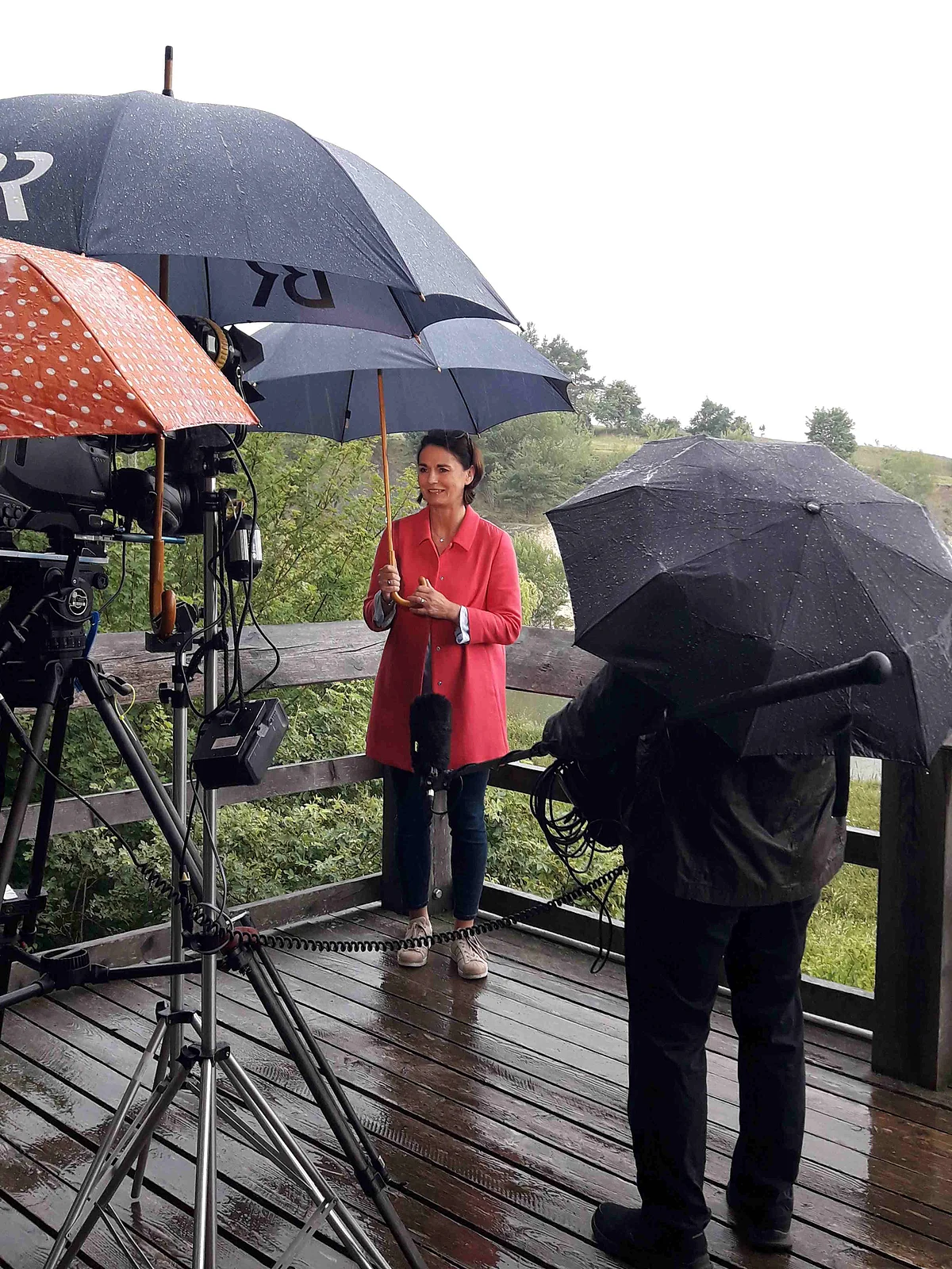 Eine Frau mit Regenschirm steht auf einer Holzplattform. Im Vordergrund sind Filmequipment und weitere Regenschirme zu sehen.