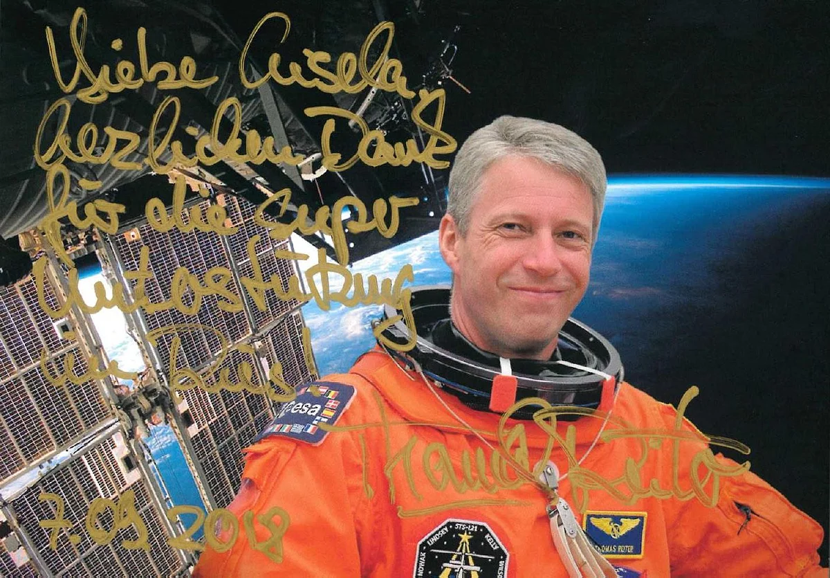 Die Autogrammkarte zeigt Thomas Reiter im orangefarbenen Astronautenanzug. Im Hintergrund ist ein Satellit und die Erde aus dem All zu sehen.