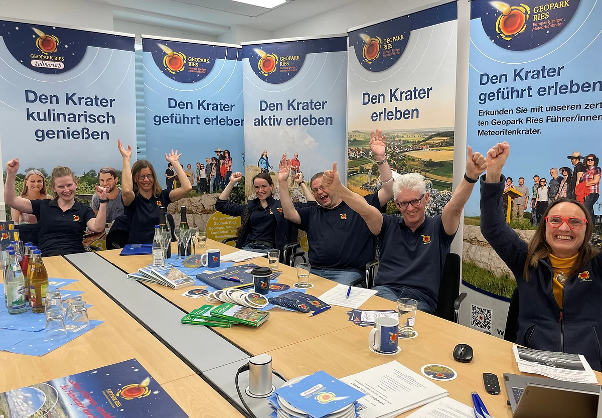 Einige Personen mit dunkelblauen Oberteilen sitzen jubelnd um einen Tisch. Im Hintergrund sind Plakate mit dem Logo des Geopark Ries zu sehen.