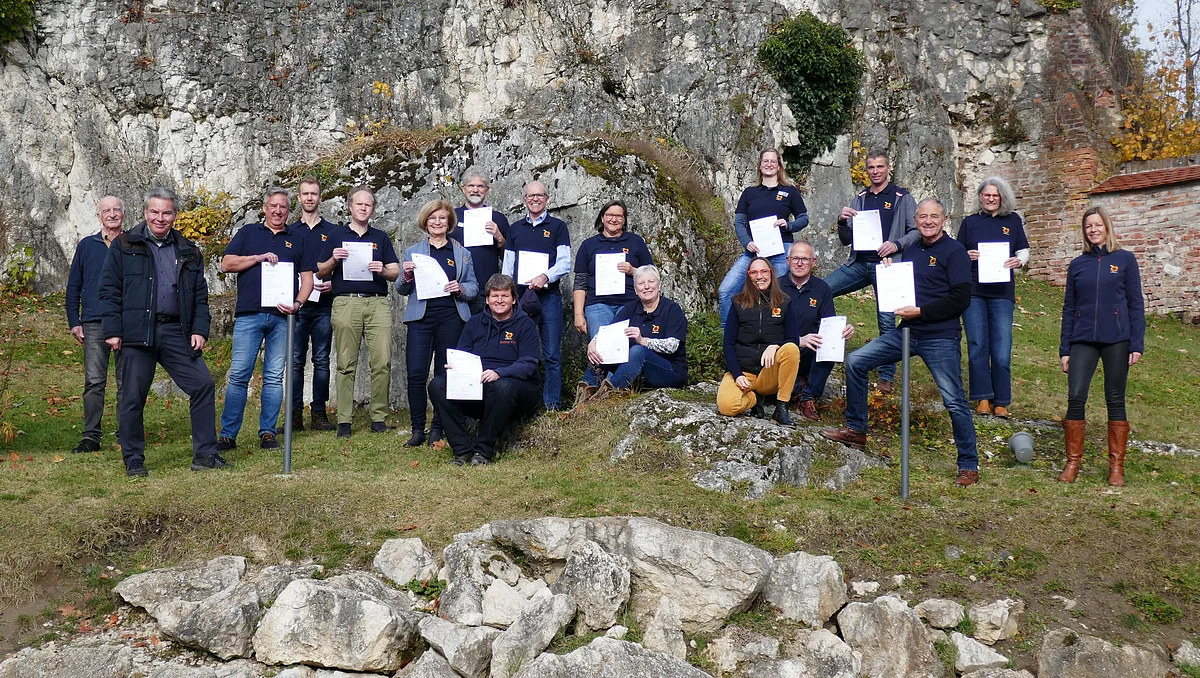 Auf einer Wiese vor einem Felsen stehen viele Personen mit dunkelblauen Oberteilen und halten eine Urkunde in den Händen.