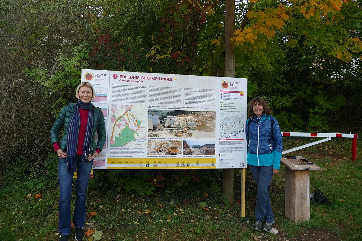 Zwei Frauen stehen neben einer großen Informationstafel mit dem Titel "Erlebnis-Geotop Lindle".