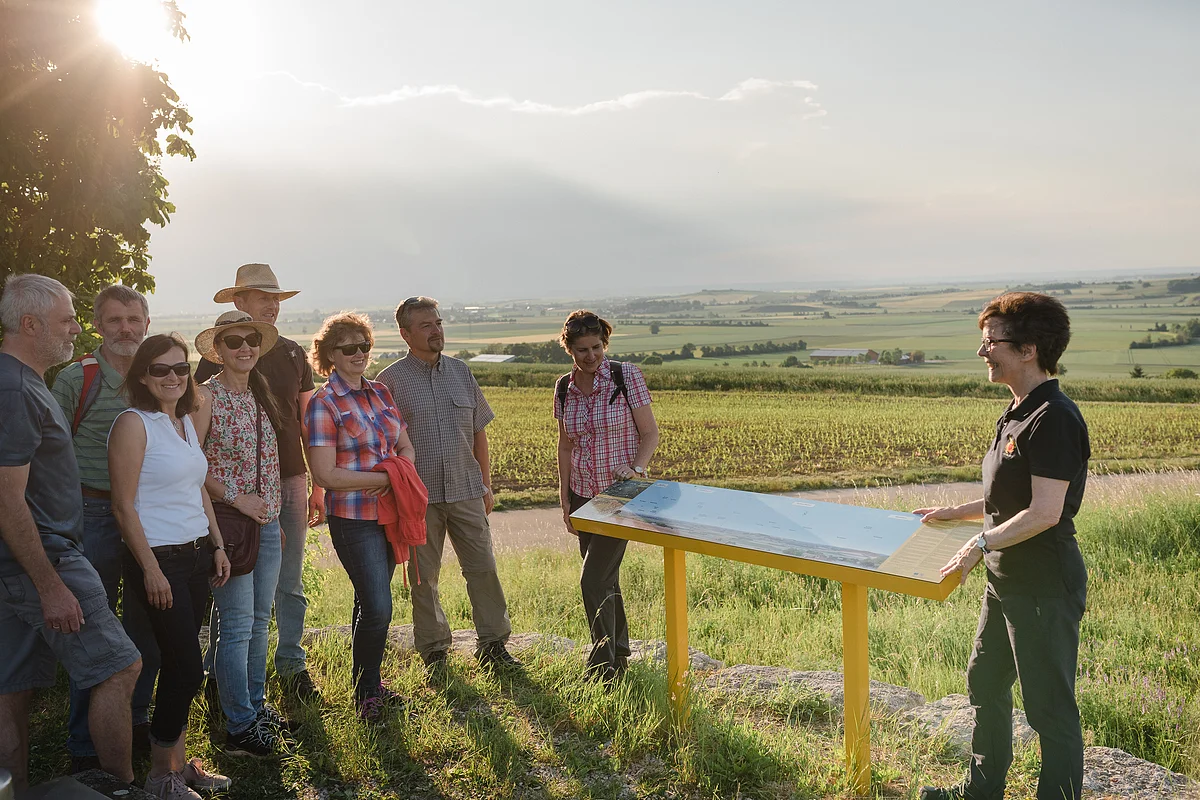 Eine Gruppe Personen steht um eine Panoramatafel, hinter der sich eine weite Landschaft mit Äckern und einzelnen Bäumen erstreckt.