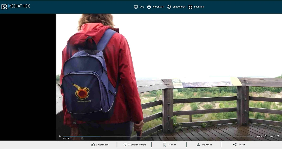 Ein Screenshot aus einem Video der BR Mediathek zeigt eine Frau mit blauem Rucksack auf einer Holzplattform.
