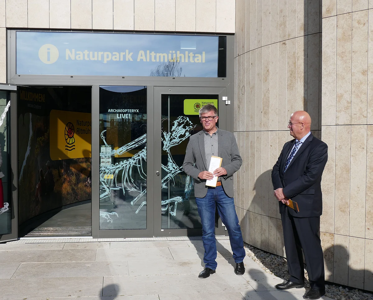 Zwei Männer stehen vor einer mit hellen Steinplatten verkleideten Fassade. In der Glasscheibe des Eingangs ist "Naturpark Altmühltal" und "Archaeopteryx live" zu lesen.