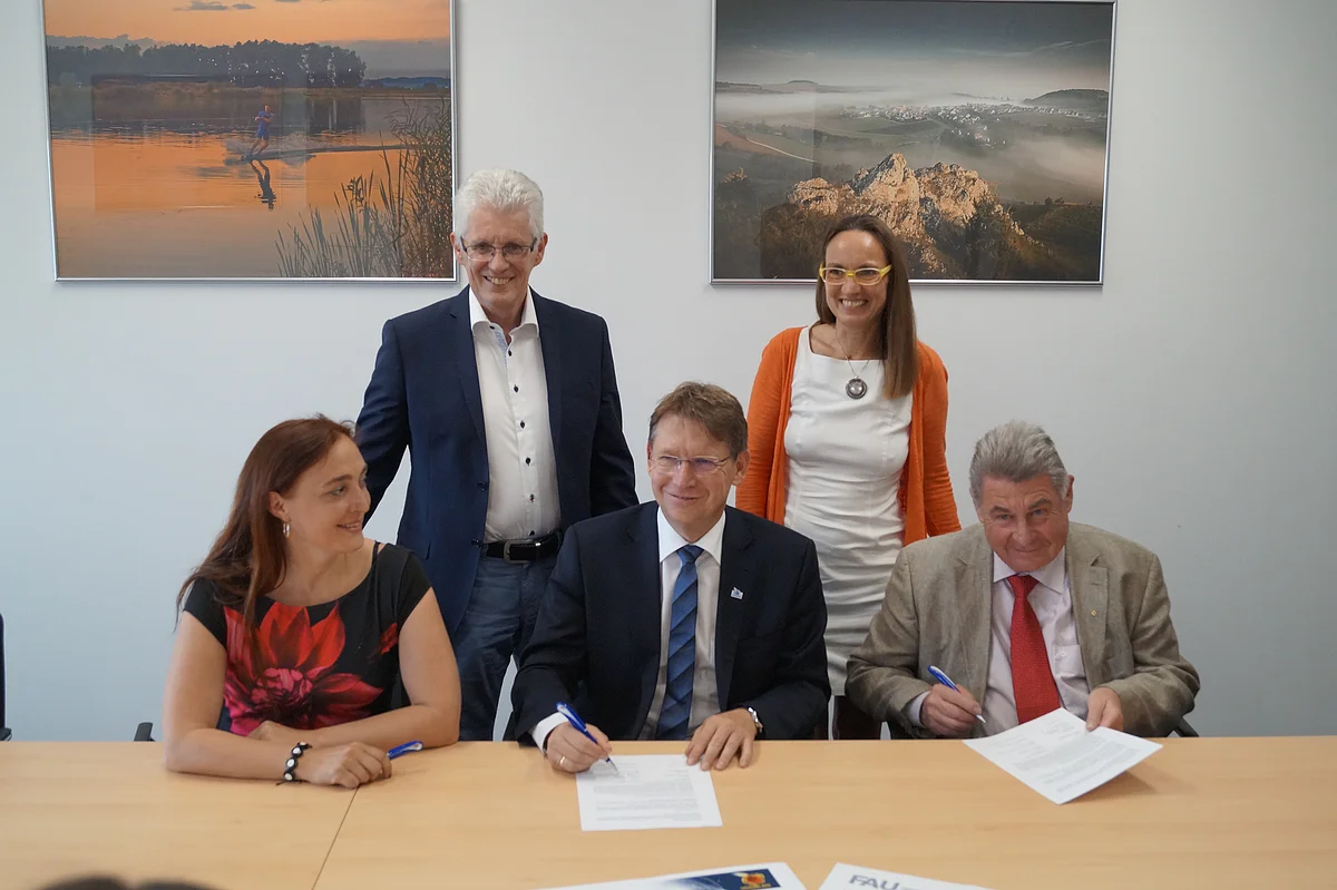 Drei Personen sitzen an einem Tisch und unterschreiben ein Blatt Papier, dahinter stehen zwei weitere Personen. An der Wand dahinter hängen großformatige Fotodrucke.