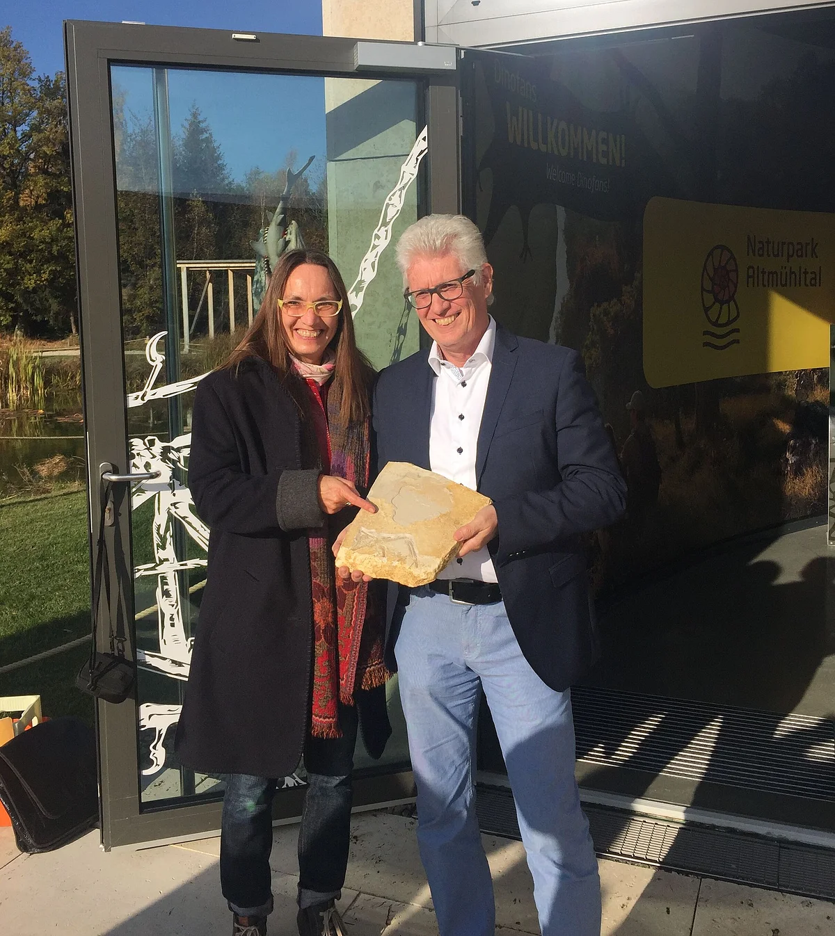 Günther Zwerger (Geschäftsführer Geopark Ries e. V.) und Heike Burkhardt (stv. Geschäftsführerin) freuen sich über den 8. Original-Archaeopterix Heike Burkhardt und Günther Zwerger halten eine Juraplatte mit versteinertem Archaeopteryx in den Händen. Im Hintergrund ist eine geöffnete Glastür zu sehen.