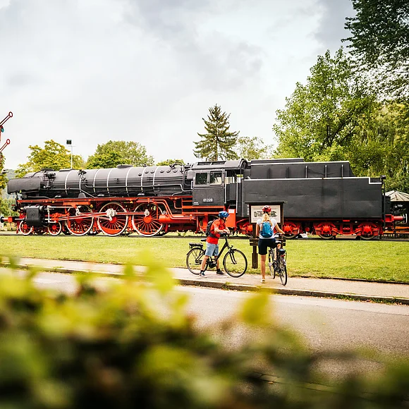 Radler vor der Eisenbahn in Treuchtlingen Dampflokomotive auf Gleis mit zwei Radfahrern davor, umgeben von Bäumen und Wiese bei bewölktem Himmel.