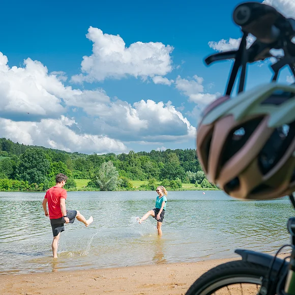 Am Hahnenkammsee Im Vordergrund stehen rechts zwei Fahrräder, dahinter liegt ein kurzer Strand an einem Seeufer. Im seichten Wasser stehen ein Mann und in eine Frau in kurzen Hosen und T-Shirts, die versuchen, sich gegenseitig nass zu spritzen. Am Ufer gegenüber liegen eine Wiese und Wald.