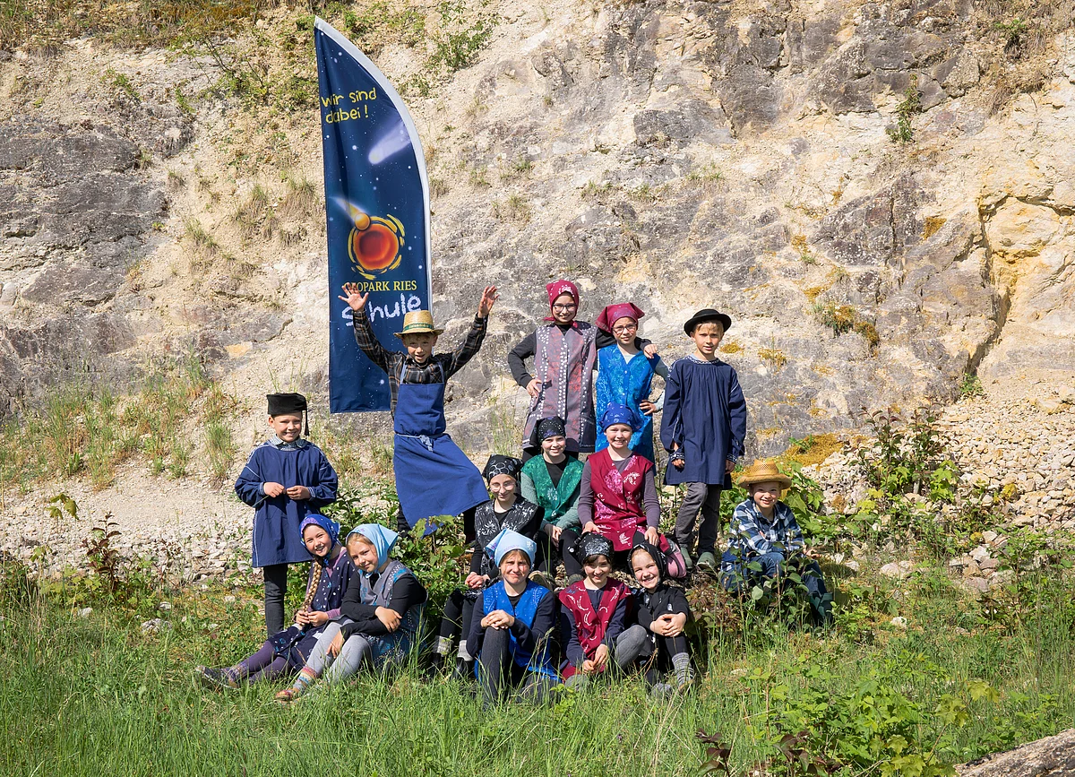Gruppenbild mit Schülerinnen und Schülern der Grundschule Mönchsdeggingen in selbst bedruckten Kittelschurzen und Bauernkitteln. Die Kinder stehen bzw. sitzen im Gras vor einer Steinbruchwand.