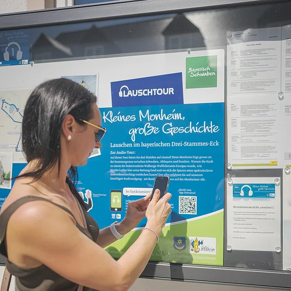 Frau mit Smartphone steht vor Infotafel zur Lauschtour in Monheim, Bayern, mit dem Titel "Kleines Monheim, große Geschichte" bei Sonnenschein. Frau hält ihr Smartphone an einen QR-Code.