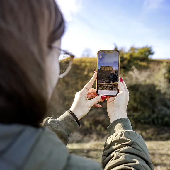 Digitaler Rundgang am Ipf Frau mit Brille und rotem Nagellack hält Smartphone mit Bild einer historischen Pfostenschlitzmauer im Freien.