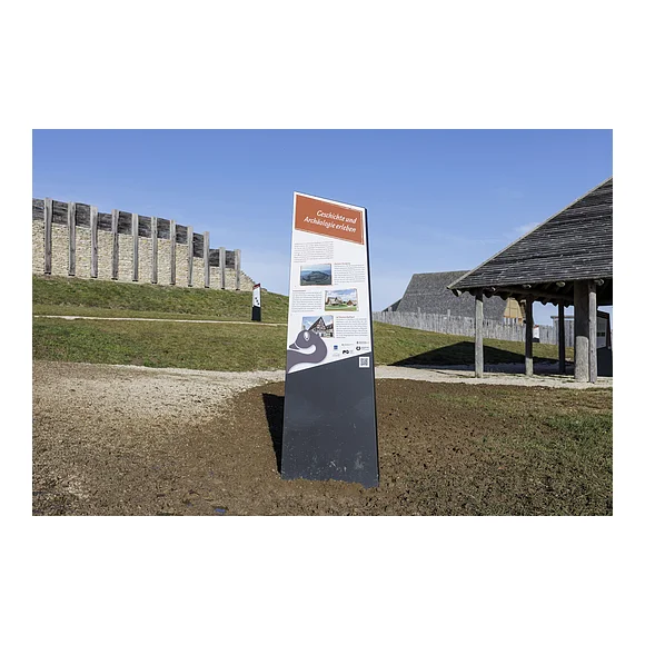 Infotafel am keltischen Freilichtmuseum mit Beschreibungstext und QR-Code zum digitalen Rundgang am Ipf Informationstafel mit dem Titel „Geschichte und Archäologie erleben“ vor einer historischen Holz- und Steinmauer bei klarem Himmel.