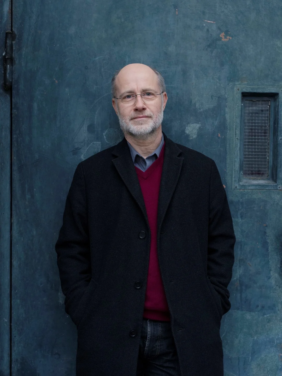 Prof. Harald Lesch steht in einem schwarzen Mantel vor einer dunklen, graublauen Wand und schaut in die Kamera.